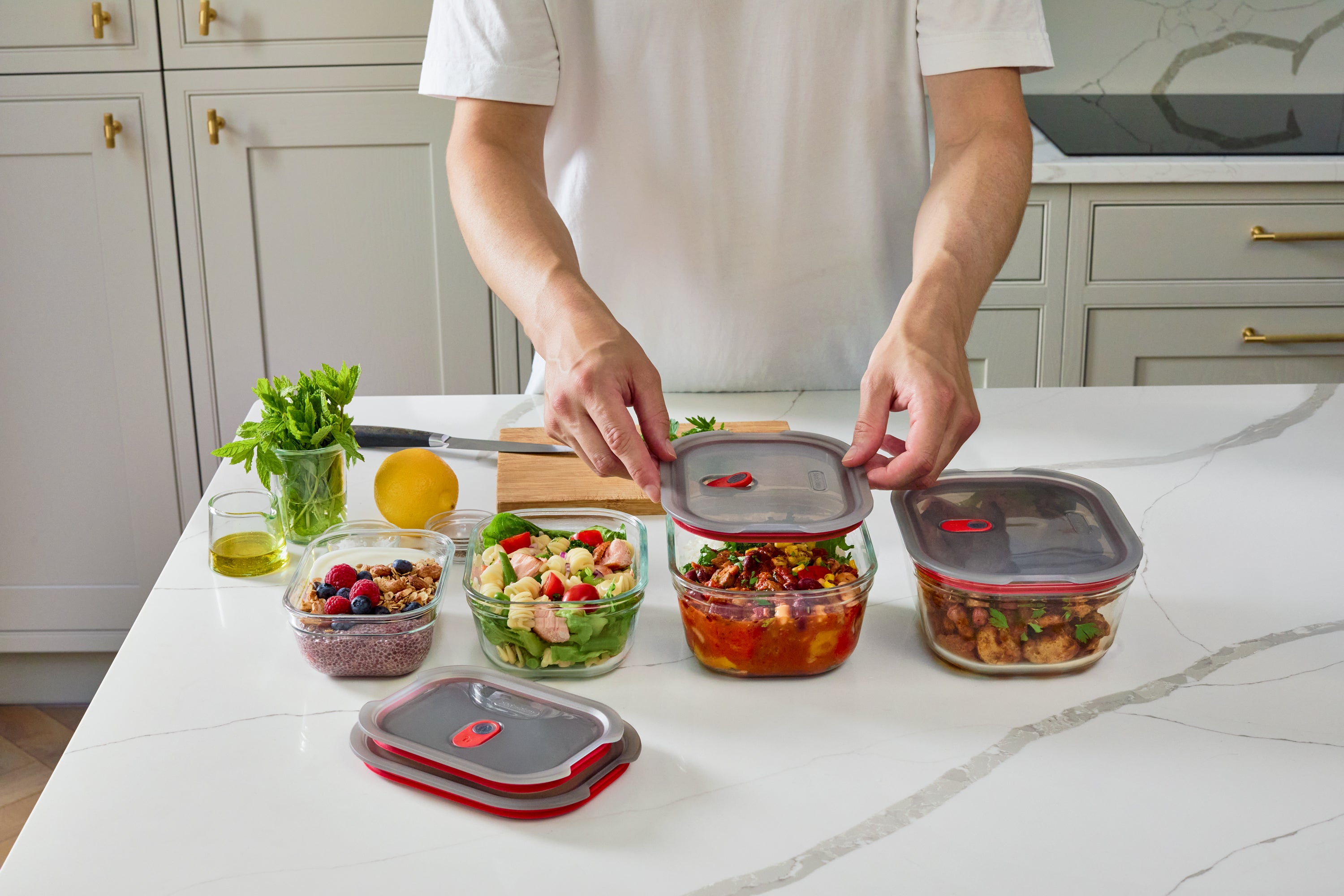 A person in a white t-shirt organizes glass food storage containers with clear lids and red silicone seals on a white marble kitchen countertop, with fresh salad, fruit, and olive oil nearby.