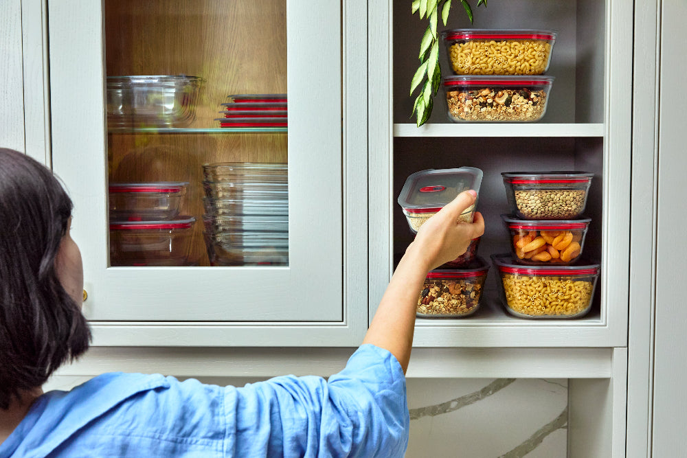 A person in a blue shirt organizes clear plastic food storage containers with red lids on a white kitchen cabinet shelf, alongside other stacked containers.