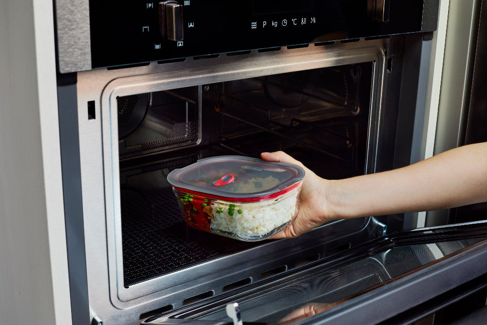 A hand placing a clear plastic food storage container with a red-trimmed lid into a stainless steel built-in oven.