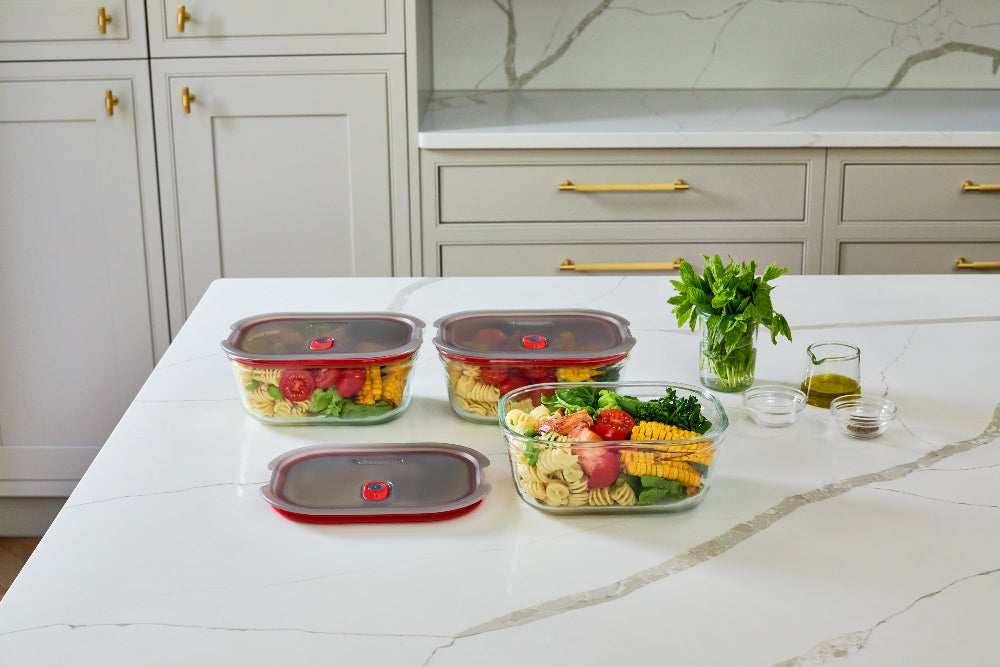 Three clear plastic food storage containers with dark lids are arranged on a white marble kitchen countertop, alongside a small glass of olive oil, a jar of herbs, and a bowl of mixed vegetables. The background shows a modern kitchen with white cabinets and gold handles.